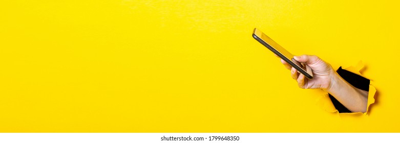 Female Hand Holds A Phone On A Torn Yellow Background. Banner.