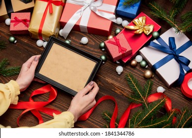 Female Hand Holds Picture Frame Near With Christmas Gifts And Pine Branches With Baubles On Wooden Table