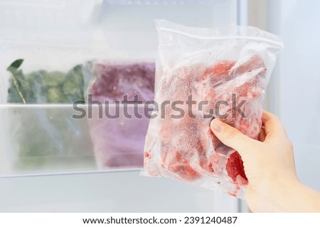 Female hand holds plastic bag with a zip fastener with frozen strawberries against the background of an open freezer with other vegetables.