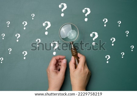 Female hand holds a plastic magnifying glass and question marks on a green background. The concept of finding an answer to questions, truth and uncertainty.