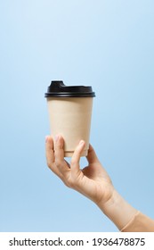 Female Hand Holds, Raised A Kraft Paper Cup Of Tea Or Coffee With Lid. Blue Background Copy Space. Vertical. Concept Of Coffee To Go, Takeaway Drinks, Cafe During Pandemic, Morning Snack, Caffeine.