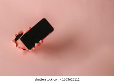 Female hand holds smartphone through round hole in pink paper. Close up, Copy space