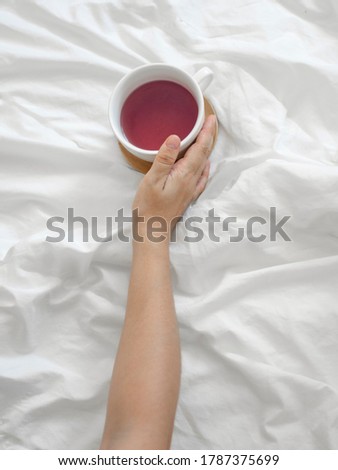 A female hand holds a white mug on a wooden stand with pink tea on the background of a white sheet