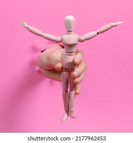 Female Hand Holds Wooden Puppet Through Torn Hole Pink Paper.