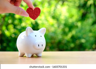 Female Hand Inserting Red Glitter Heart Into White Ceramic Piggy Bank On Wood Table With Blurred Bokeh Outdoor Green Nature Background. Concept For Money Savings, Retirement Plan, Or Fundraising.