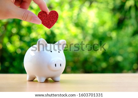 Female hand inserting red glitter heart into white ceramic piggy bank on wood table with blurred bokeh outdoor green nature background. Concept for money savings, retirement plan, or fundraising.