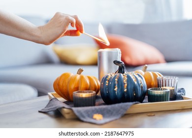 Female hand lighting a candle with a match in autumn cozy composition for hygge home mood. Pumpkins and candles on the tray with gray napkin on the coffee table in the living room. Selective focus.