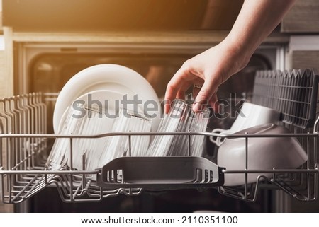 Female hand loading dished, empty out or unloading dishwasher with utensils. Kitchen appliances, lifestyle view. Woman puts a plate in the dishwasher or takes from it. Housewife does her housework 