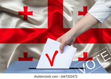 Voter’s Female Hand Lowers The Ballot In The Ballot Box Against The Background Of The National Flag Of Georgia, Concept Of State Elections