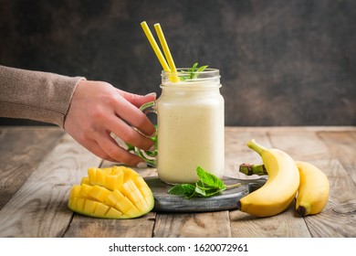 Female Hand With Mango And Banana Smoothie On A Rustic Background. Healthy Food, Detox Diet. Copy Space.