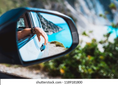 Female hand mirrored in the car side view mirror. Blue mediterranean sea and white rocks in background