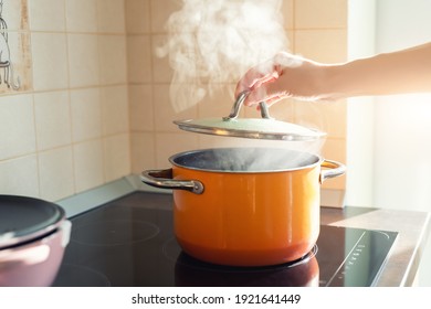 Female hand open lid of enamel steel cooking pan on electric hob with boiling water or soup and scenic vapor steam backlit by warm sunlight at kitchen. Kitchenware utensil and tool at home background - Shutterstock ID 1921641449