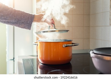 Female hand open lid of enamel steel cooking pan on electric hob with boiling water or soup and scenic vapor steam backlit by warm sunlight at kitchen. Kitchenware utensil and tool at home background - Shutterstock ID 1922416616