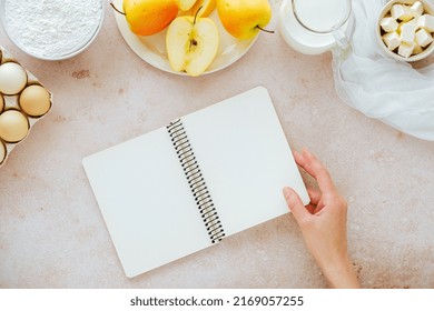 Female Hand Opening Cook Book With Ingredients Flour, Milk, Eggs, Apples, Butter. Recipes For Autumn Vegan Baking. Flat Lay, Top View. High Quality Photo