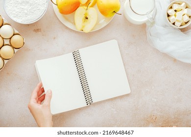 Female Hand Opening Cook Book With Ingredients Flour, Milk, Eggs, Apples, Butter. Recipes For Autumn Vegan Baking. Flat Lay, Top View. High Quality Photo