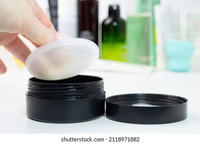 A female hand opens a protective cap in a plastic jar of cream, gel, patches on the table with other cosmetics