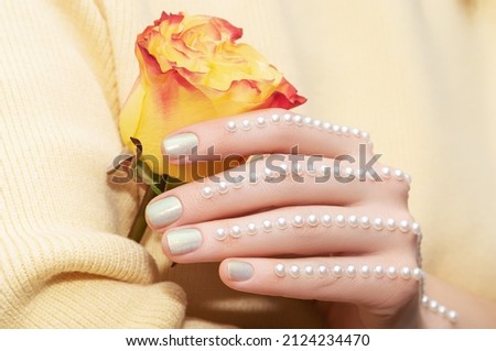 Female hand with pearlescent nail design. Glitter pearlescent nail polish manicure with gems nail art. Model hand with pearlescent manicure hold yellow rose flower on yellow background. Copy space.