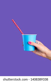 Female Hand With Pink Manicure Holding Blue Paper Cup Template For Soda Or Cold Beverage With Red Drinking Straw, Isolated On Lilac Blue Background. Vertical Shot.