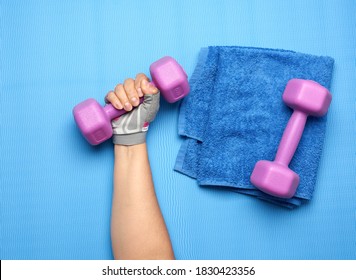 Female Hand In A Pink Sports Glove Holds A Purple One Kilogram Dumbbell On A Blue Background