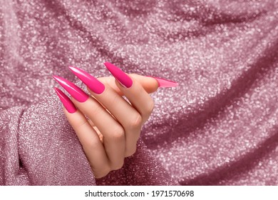 Female hand with pink stiletto nail design. Long nail polish manicure. Woman hand on glitter pink fabric background.