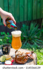 A Female Hand Pouring Beer From The Bottle Into A Glass Set On The Wooden Stamp In The Garden