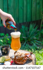 A Female Hand Pouring Beer From The Bottle Into A Glass Set On The Wooden Stamp In The Garden