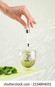 Female hand pours liquid chlorophyll into a glass of water with a dropper. Liquid chlorophyl and fresh herbs on the green table. Green background, natural sunlight, vertical. Superfood and healthy.