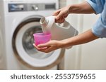 female hand pours liquid transparent laundry detergent or conditioner into plastic cap against blurred washing machine.