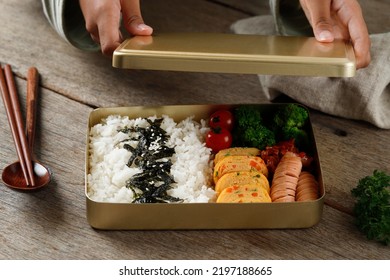 Female Hand Preparing Close The Lid Box Of Dosirak Or Korean Bento Box, Served On Gold  Metal Lunchbox To Keep Food Warm. Korean Popular Packed Lunch 