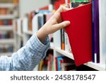 Female hand pulling book from bookshelf in public library in university, college or high school. Woman student take novel from bookcase in bookshop store, soft focus. Education and literature concept