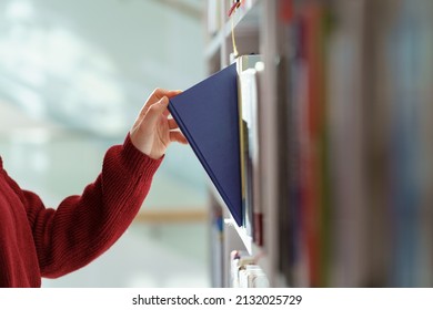Female hand pulling book from bookshelf in public library in university, college or high school. Woman student take novel from bookcase in bookshop store, soft focus. Education and literature concept