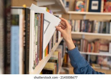 Female hand pulling book from bookshelf in public library in university, college or high school. Woman student take novel from bookcase in bookshop store, soft focus. Education and literature concept