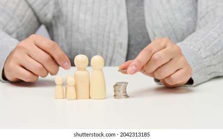 Female Hand Puts Coins In A Pile And Wooden Figurines Of A Family On A White Table. Economy And Investment Concept, Budget Planning. Receiving Subsidies From The State, Tax