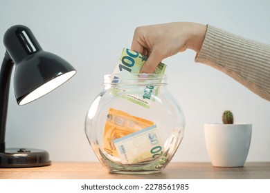 Female hand putting 100 euro banknote into glass jar, table lamp and cartus nearby. Concept of savings, collecting money for a dream - Powered by Shutterstock