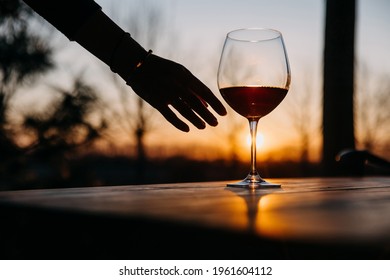 Female hand reaching a glass of red wine on a wooden table, at sunset.