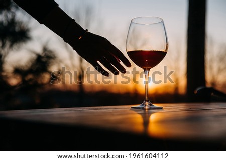 Female hand reaching a glass of red wine on a wooden table, at sunset.