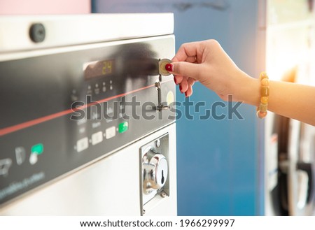 Female hand ready to insert a token into the slot of vintage chrome coin on the washing machine. Asian woman hand putting coin in washer machine for operated, inside laundrette, closeup