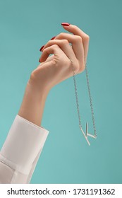 A female hand with red manicure is holding a silver chain with a pendant in the shape of a letter K. The necklace is hanging from the wrist on the blue backdrop.