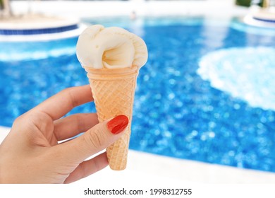 Female Hand With Red Manicure Holds Yellow Ice Cream On A Hot Day On Background Of A Blue Pool. The Concept Of Leisure, Summer Vacation. Copy Space.