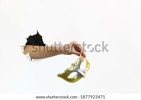 A female hand with a red manicure through torn white paper holds a carnival mask. Hand through torn white paper