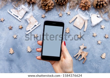 Female hand with red nail polish holding a blank smart mobile phone above grey table with Christmas wooden decoration and pine cones