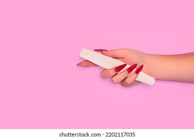Female hand with red stiletto nail design. Glitter red nail polish manicure. Female hand with stiletto nails hold white nail file on pink background. Copy space. Plce for text.