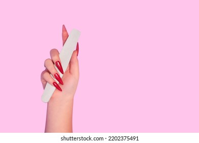 Female hand with red stiletto nail design. Glitter red nail polish manicure. Female hand with stiletto nails hold white nail file on pink background. Copy space. Plce for text.