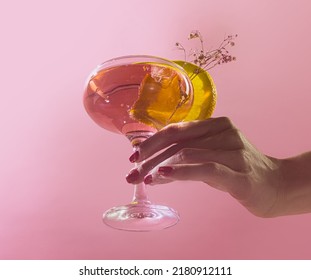 Female Hand Rising Glass Of Delicious Alcoholic Cocktail With Rose Water And Sparkling Wine Isolated Over Pink Background. Concept Of Cocktails, Taste, Party, Mix. Copy Space For Ad. Retro Style