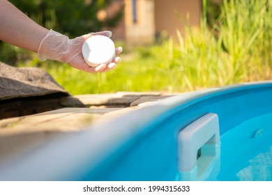 Female hand in rubber glove holds white chlorine pill over skimmer of swimming pool with blue water. Cleaning, disinfection of water in swimming pool.