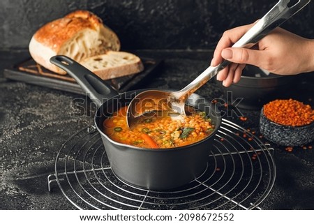 Female hand with saucepan of hot lentil soup and ladle on dark background