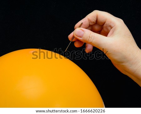 In a female hand a sewing needle. The needle is above the surface of an orange balloon. Concept-criticism, debunking of false authorities