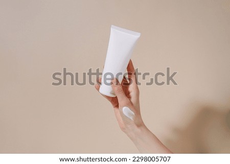Female hand with smear holding blank mockup tube of cream on beige isolated background. Daily skincare and body care routine. Image for your design