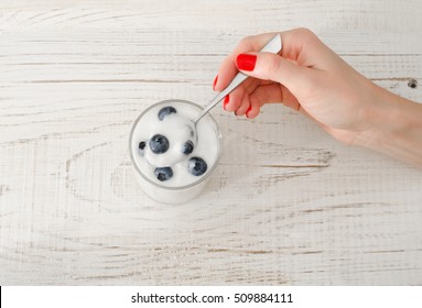 Female Hand With A Spoon Yogurt With Blueberries On White Wooden Table, Top View