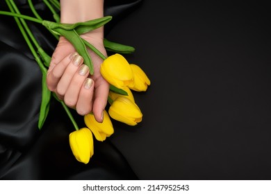Female hand with spring nail design. Glitter golden nail polish manicure. Female model hand with perfect manicure hold yellow tulip flowers on black fabric background